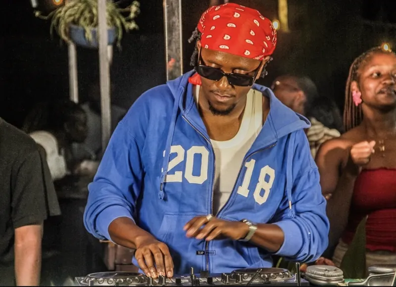 DJ in a red paisley bandana and blue hoodie mixing at night while the crowd dances behind