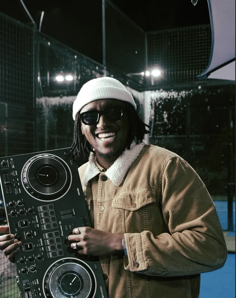 DJ in a white beanie and tan corduroy jacket smiling, holding a Pioneer DJ controller under floodlights