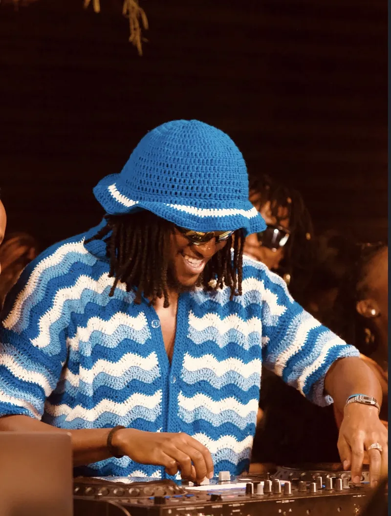 DJ grinning at the decks in a blue-and-white crocheted wave bucket hat, warm amber light